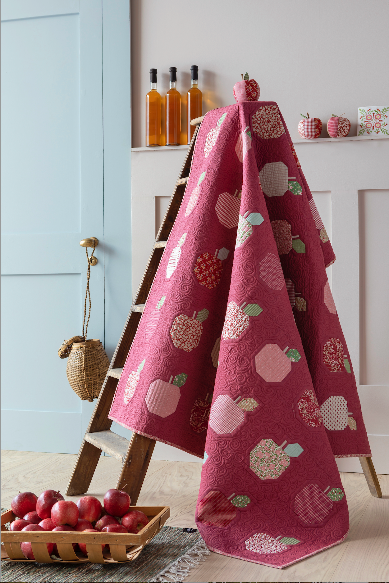 A red quilt with a pattern of stylized apples in various shades, draped over a wooden ladder in a styled kitchen setting.
Photo by Tildafabrics.com [additional_image_link]