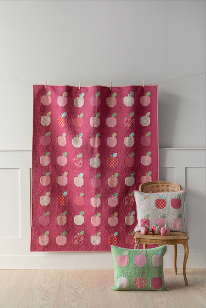 A quilt kit display featuring a red and green applique quilt with a pattern of apples, accompanied by pillows with a similar design, hung on a wall in a room with wooden flooring and a white background.
Photo by Tildafabrics.com [image_link]
