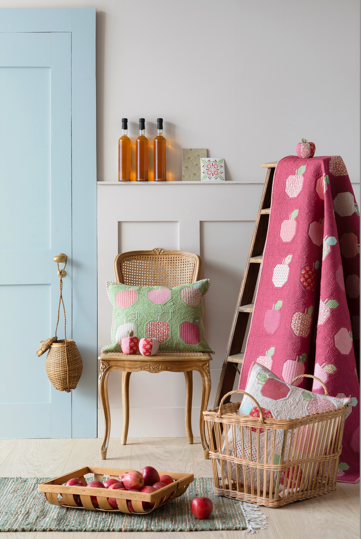 A styled interior scene with a pink blanket draped over a wooden ladder, featuring a white apple pattern. A small wicker chair holds a green pillow with pink apple motifs, next to a wicker basket with a matching pillow.
Photo by Tildafabrics.com [additional_image_link]