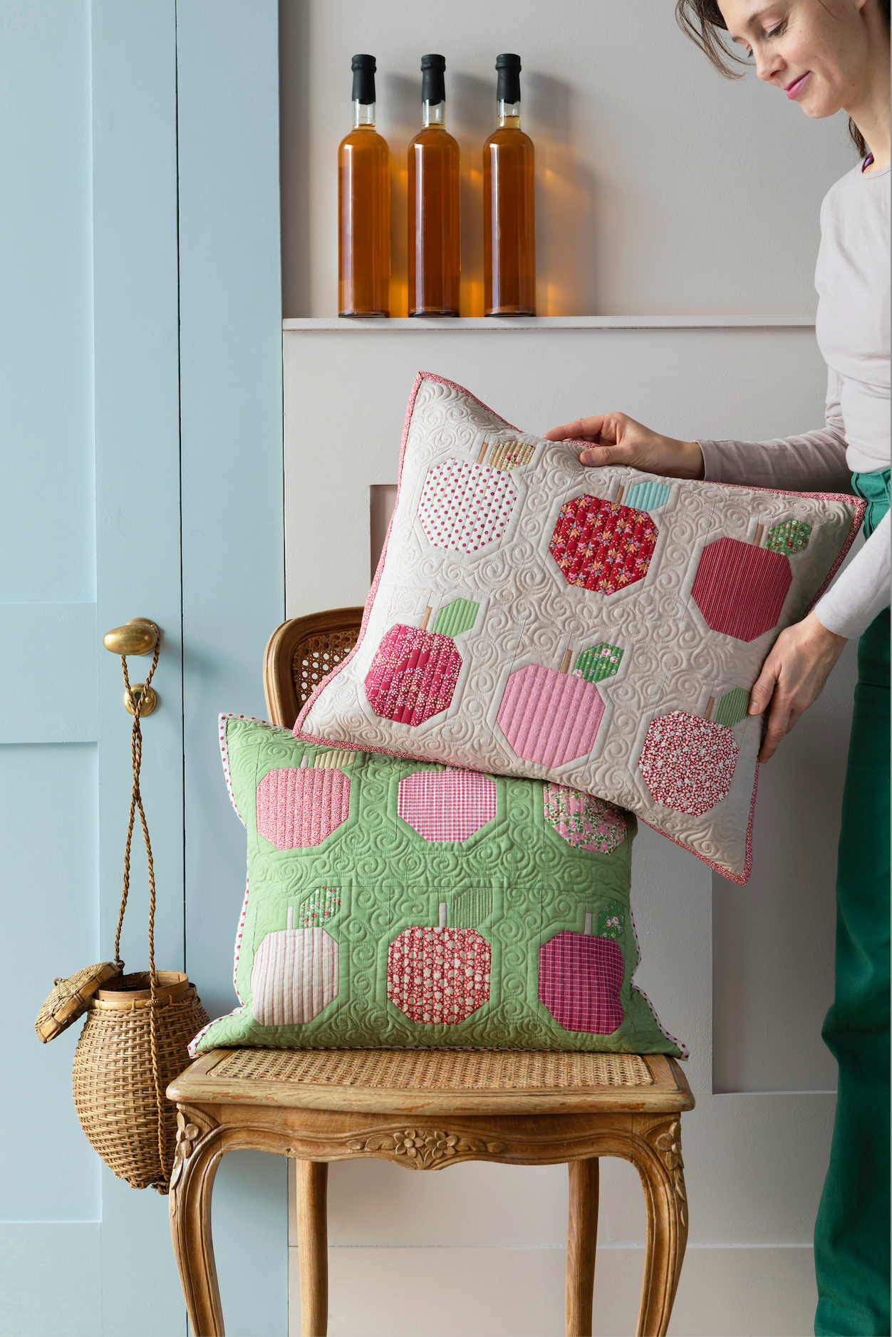 A woman in a white top and green skirt holds a quilted throw pillow with a pink and red apple pattern, standing beside another matching pillow on a wooden chair in a styled room.
Photo by Tildafabrics.com [additional_image_link]