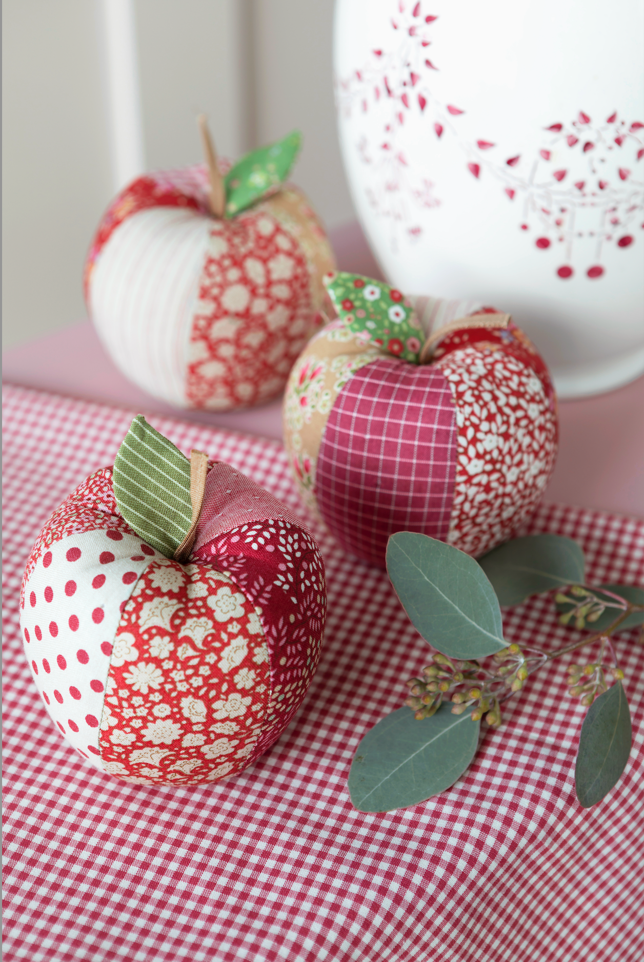 Three handmade fabric apples with red and white patterned patches and green leaves, arranged on a red-and-white gingham tablecloth with a white decorative vase in the background.
Photo by Tildafabrics.com [additional_image_link]