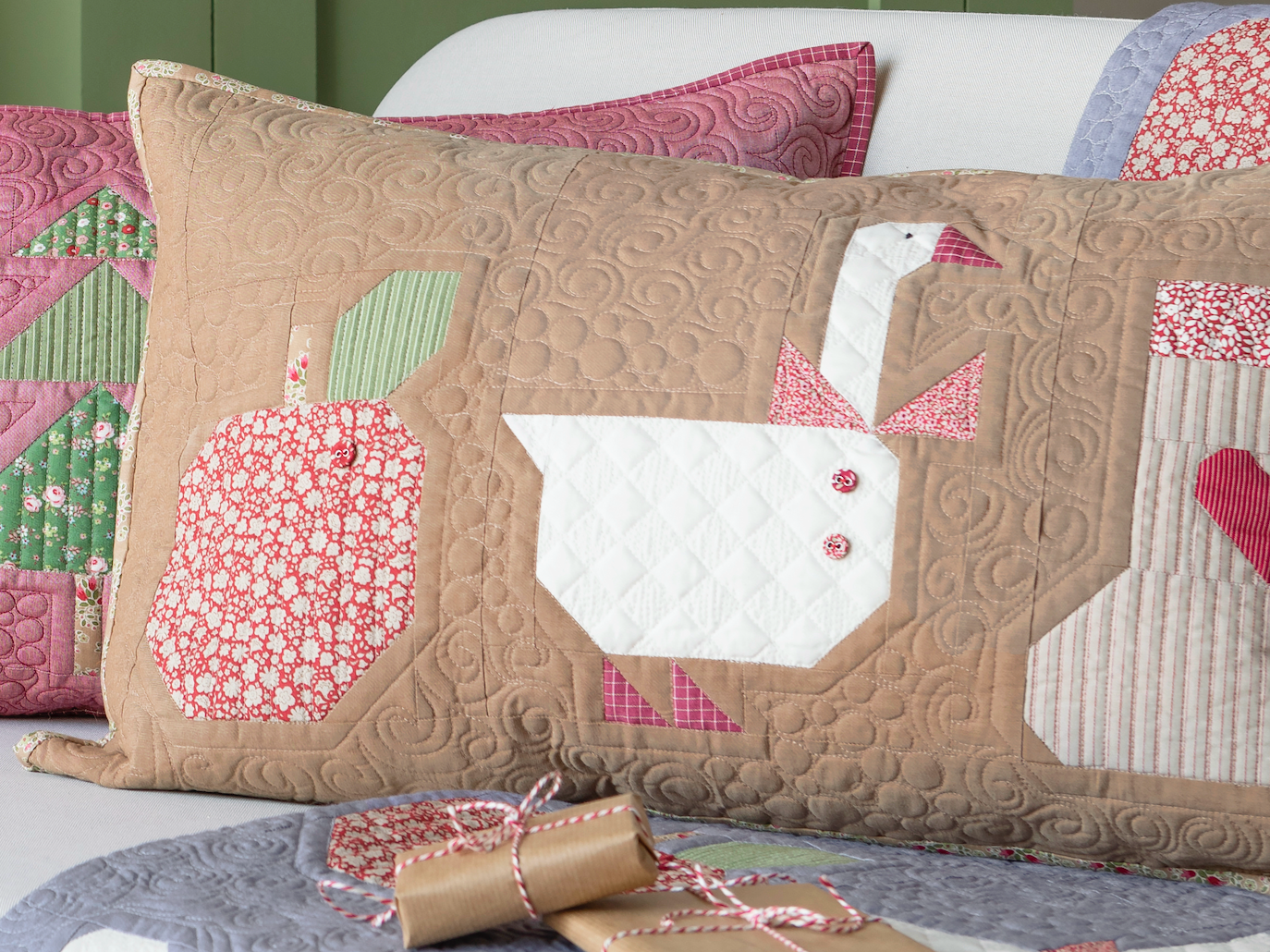 A quilted pillow with a brown chambray background featuring various Christmas-themed fabric patches, including an apple, a goose, and Christmas trees. The pillow is part of a quilting kit and is presented with its pattern and a Tilda button.
Photo by Tildafabrics.com [image_link]