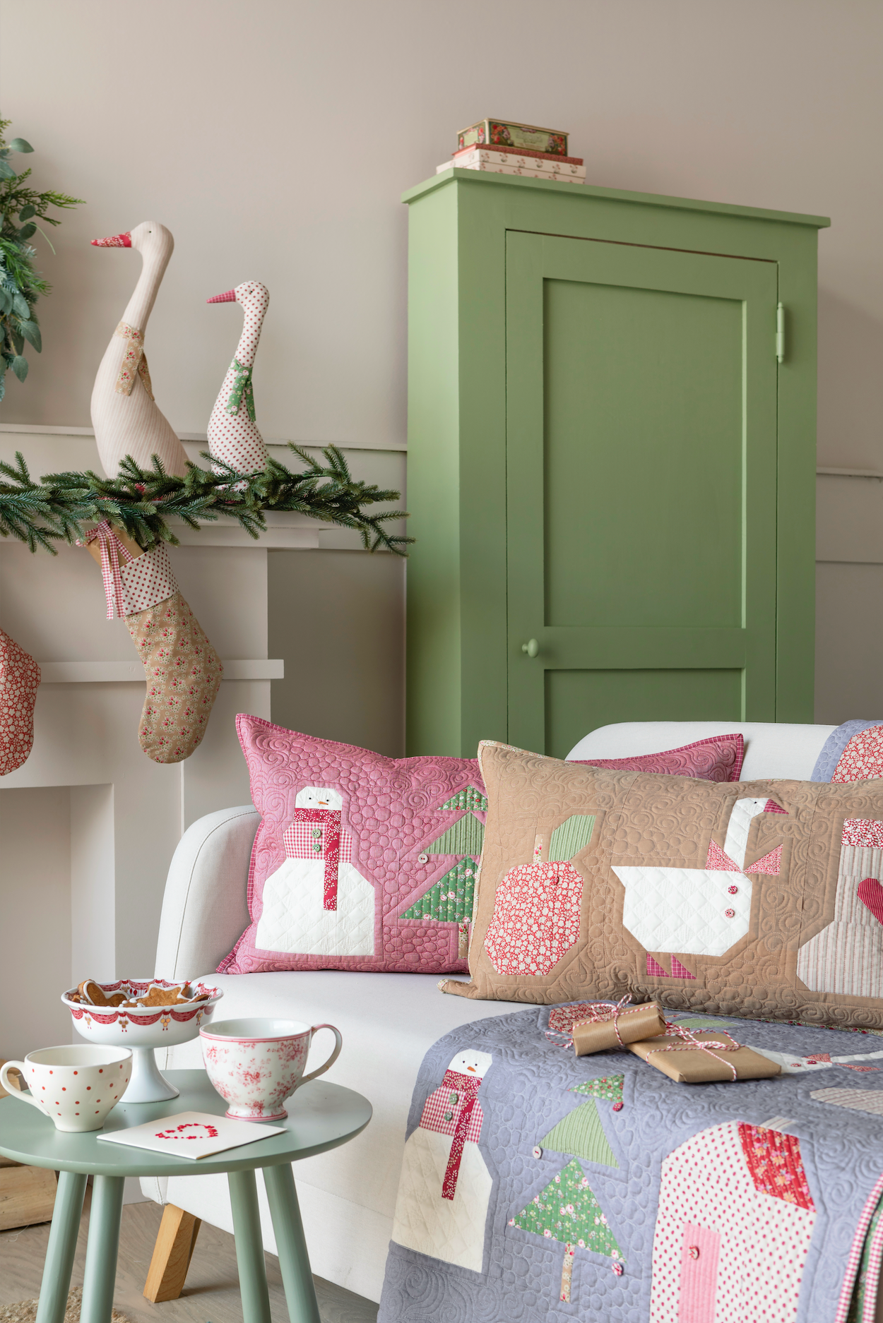 A cozy living room scene featuring a white sofa styled with pink and beige winter-themed quilted throw pillows, a matching blue quilted blanket with house and tree motifs, a small mint green side table, and a tall sage green cabinet in the background.
Photo by Tildafabrics.com [additional_image_link]