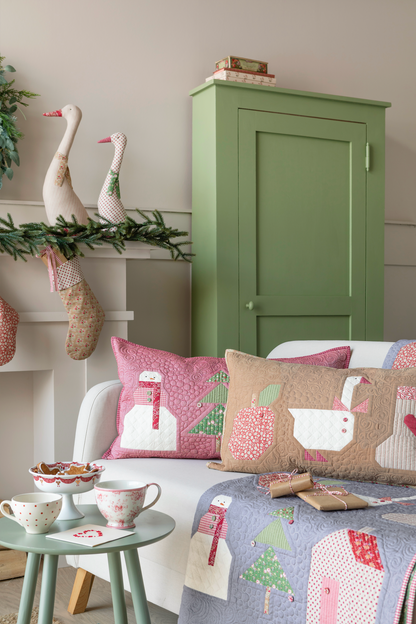 A cozy living room scene featuring a white sofa styled with pink and beige winter-themed quilted throw pillows, a matching blue quilted blanket with house and tree motifs, a small mint green side table, and a tall sage green cabinet in the background.
Photo by Tildafabrics.com [additional_image_link]