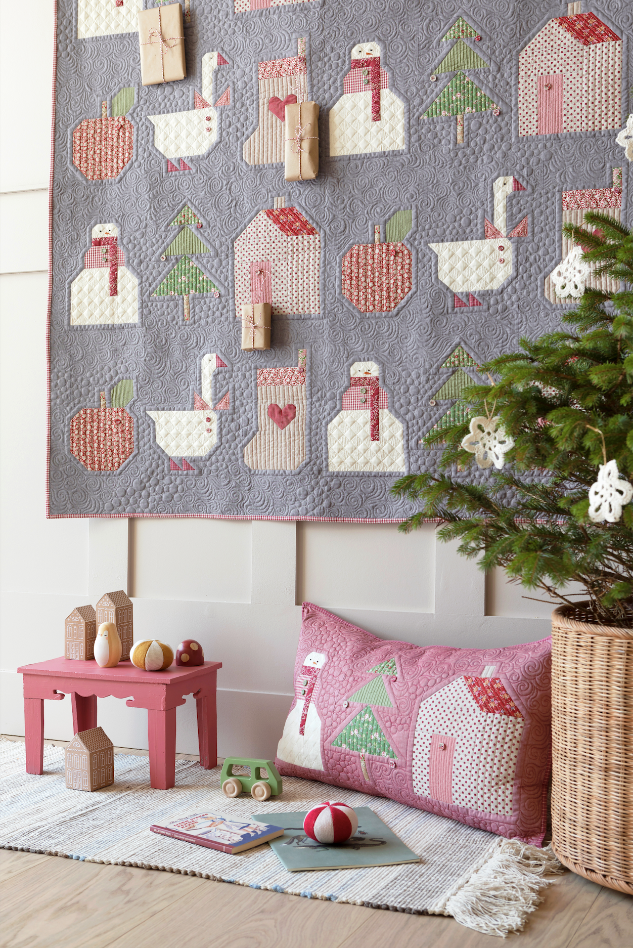 A cozy nursery or playroom scene featuring a large gray quilted wall hanging and a matching pink throw pillow with whimsical applique motifs including houses, trees, and animals. The room includes a small pink table, wooden blocks, a rug, and a potted evergreen tree.
Photo by Tildafabrics.com [additional_image_link]