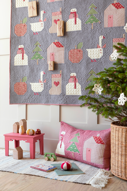 A cozy nursery or playroom scene featuring a large gray quilted wall hanging and a matching pink throw pillow with whimsical applique motifs including houses, trees, and animals. The room includes a small pink table, wooden blocks, a rug, and a potted evergreen tree.
Photo by Tildafabrics.com [additional_image_link]