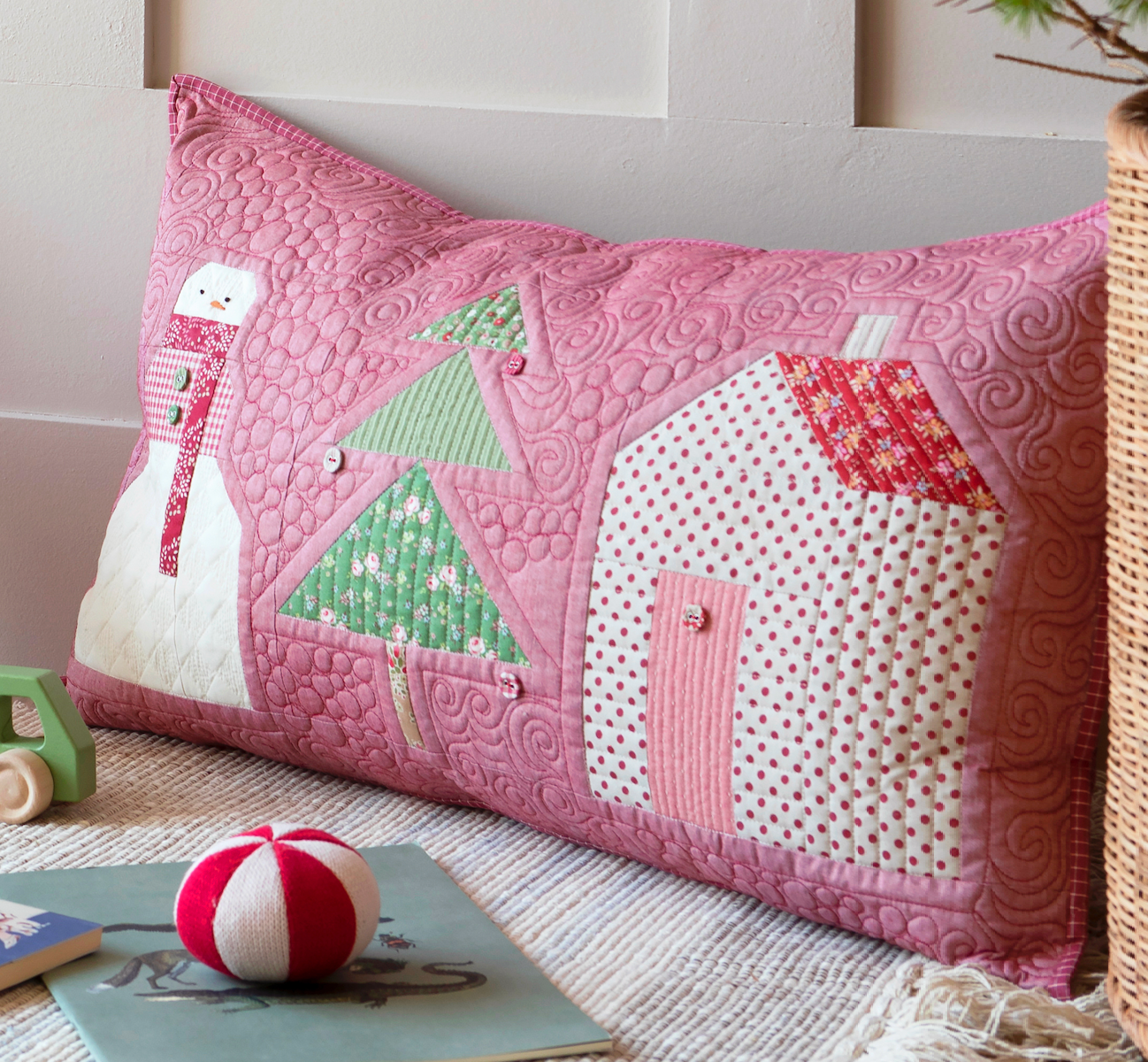 A pink quilted lumbar pillow with a patchwork winter design, featuring a snowman, green trees, and houses, styled on a bed with toys and a basket.
Photo by Tildafabrics.com [additional_image_link]
