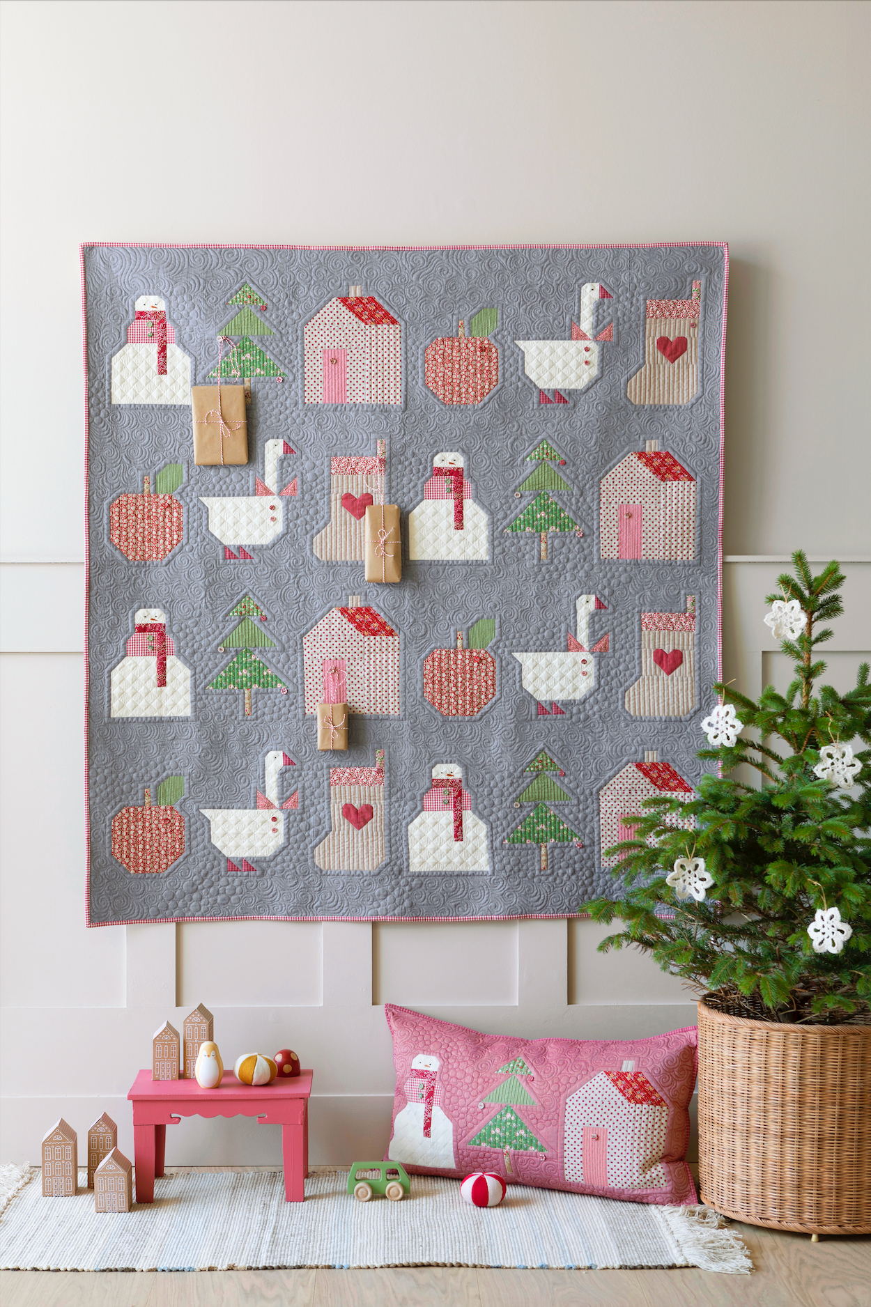 A square quilted wall hanging or blanket with a gray background and a repeating pattern of white swans, pink houses, red apples, green trees, and pink stockings, styled in a nursery-like room with a matching pillow, small toys, and a potted plant.
Photo by Tildafabrics.com [additional_image_link]