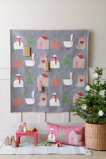 A square quilted wall hanging or blanket with a gray background and a repeating pattern of white swans, pink houses, red apples, green trees, and pink stockings, styled in a nursery-like room with a matching pillow, small toys, and a potted plant.
Photo by Tildafabrics.com [additional_image_link]