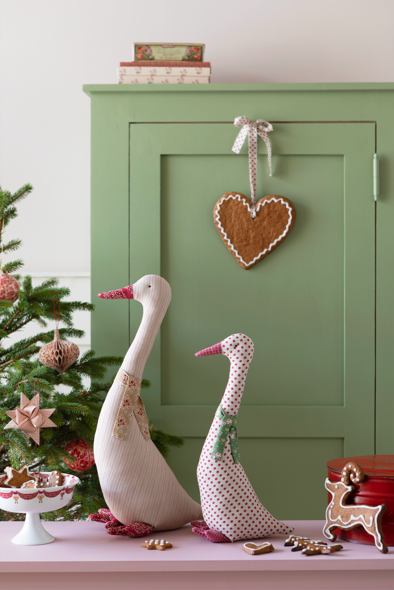 A styled holiday scene with two decorative goose figurines on a pink surface in front of a sage green cabinet. A gingerbread heart hangs from the cabinet, and a small Christmas tree, bowls, and gingerbread cookies are visible.
Photo by Tildafabrics.com [additional_image_link]