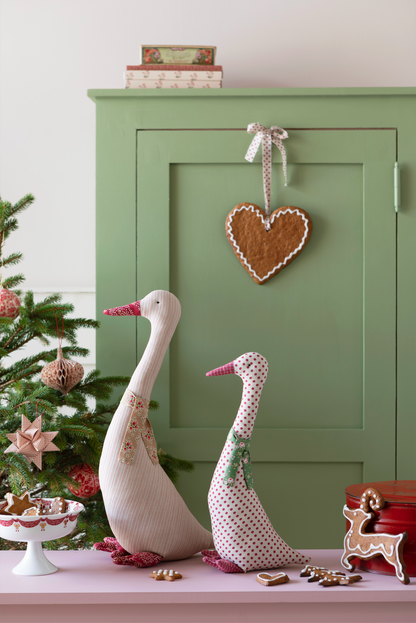 A styled holiday scene with two decorative goose figurines on a pink surface in front of a sage green cabinet. A gingerbread heart hangs from the cabinet, and a small Christmas tree, bowls, and gingerbread cookies are visible.
Photo by Tildafabrics.com [additional_image_link]