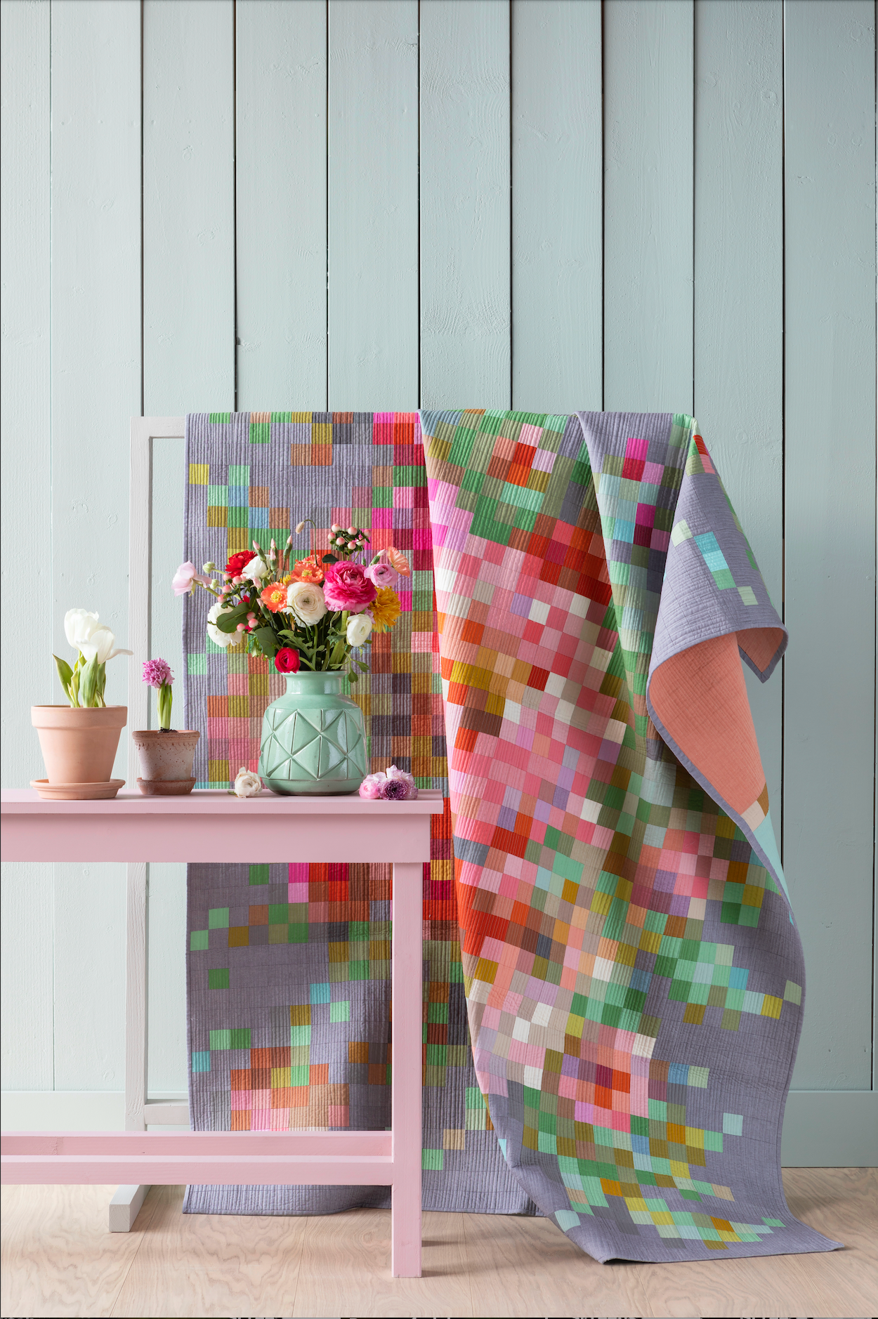 A colorful quilted blanket with a pixelated floral pattern draped over a white stand next to a pink side table with potted plants and a vase of flowers, set against a light blue paneled wall.
Photo by Tildafabrics.com [additional_image_link]