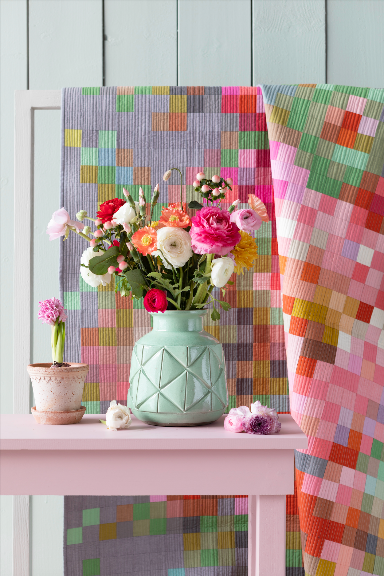 A pastel pink table styled with a mint green geometric ceramic vase holding a colorful bouquet of flowers, set against a backdrop of folded quilted fabrics in a patchwork pattern of pink, green, yellow, and blue.
Photo by Tildafabrics.com [additional_image_link]