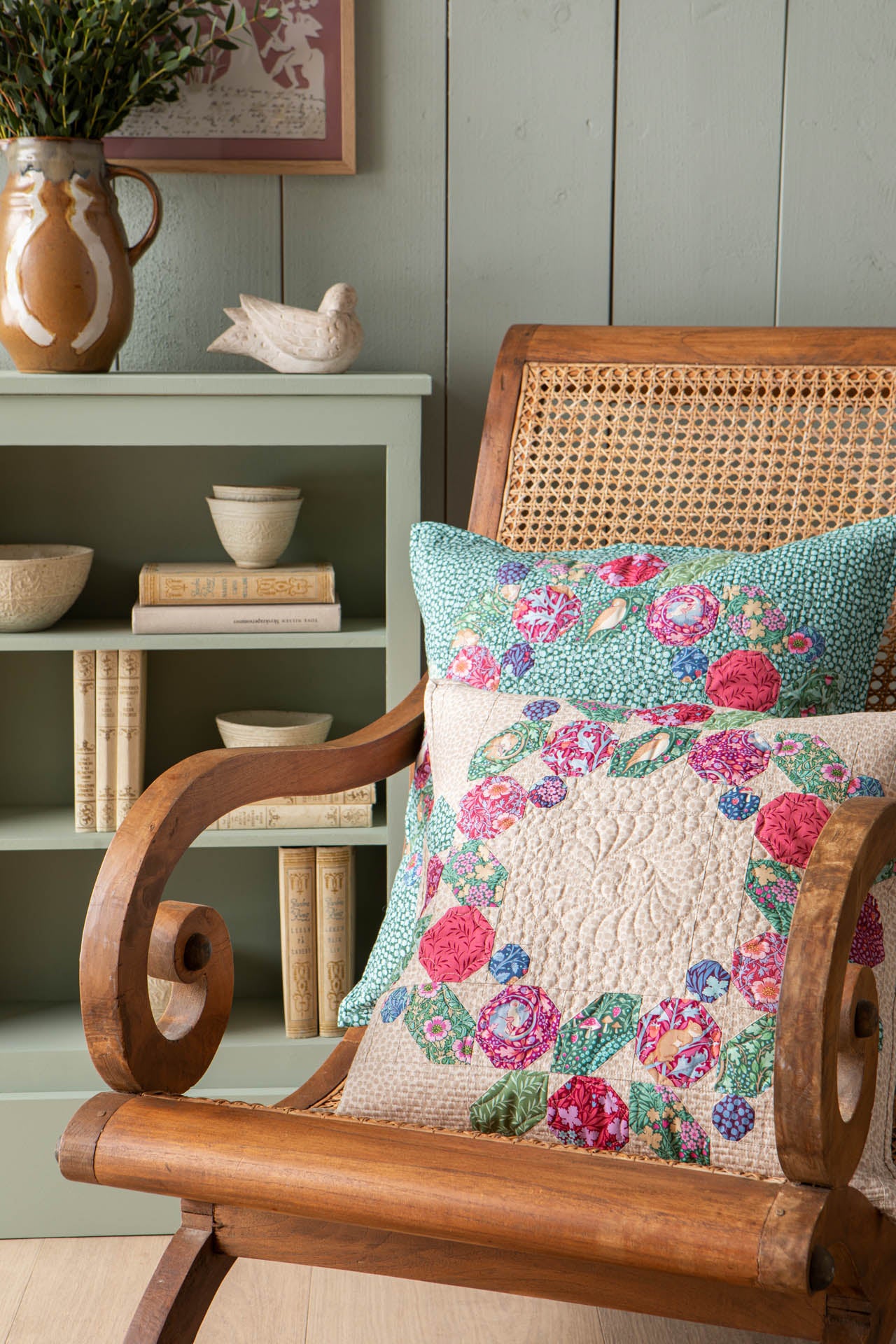 A cozy interior scene showing a cream quilt and matching pillow with a bright pink, teal, and green floral print, styled on a cane chair beside a small decorated tree and a mint green side table.
Photo by Tildafabrics.com [image_link]