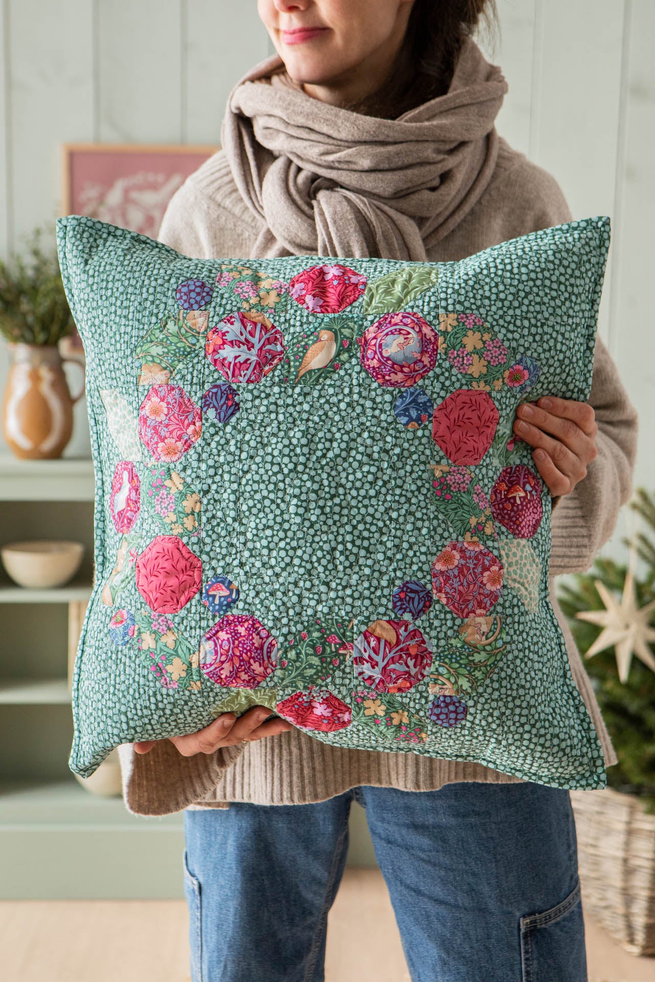 A person wearing a beige knit sweater and scarf holds a square decorative pillow. The pillow has a mint green background with a dense small floral pattern and a border of larger pink, red, purple, and blue flowers.
Photo by Tildafabrics.com [additional_image_link]