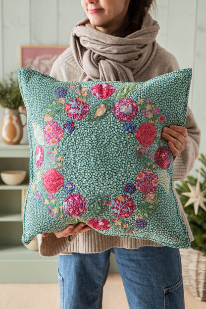 A person wearing a beige knit sweater and scarf holds a square decorative pillow. The pillow has a mint green background with a dense small floral pattern and a border of larger pink, red, purple, and blue flowers.
Photo by Tildafabrics.com [additional_image_link]