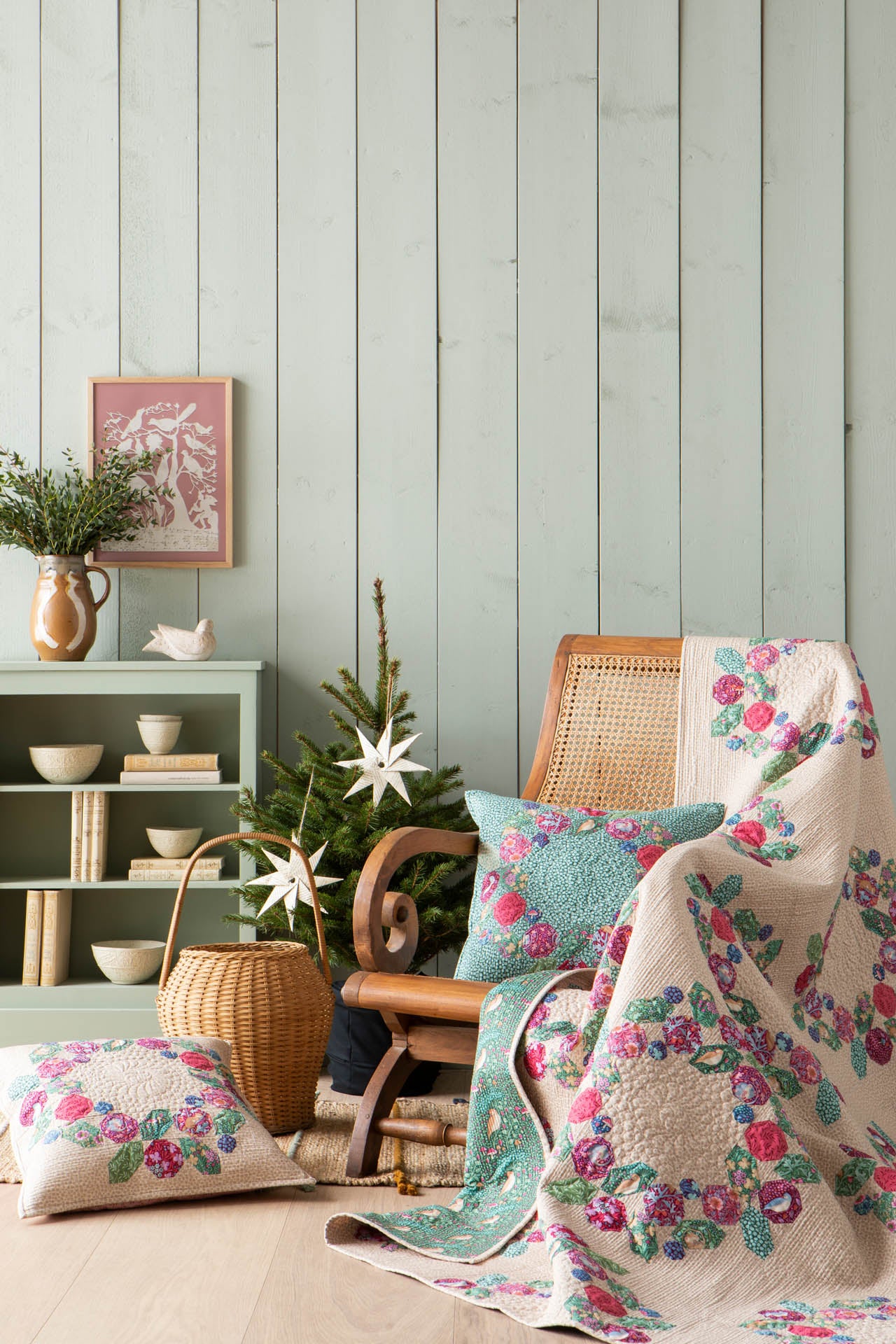 A cozy interior scene with a floral quilt draped over a wooden chair, a matching pillow on the floor, a teal accent pillow, a small tree, and a pale green shelving unit against a light wood-paneled wall.
Photo by Tildafabrics.com [additional_image_link]