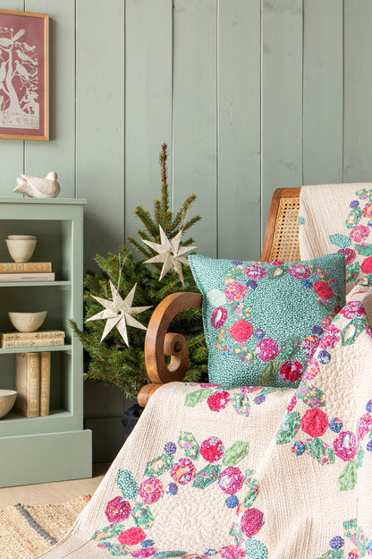 A cozy interior scene showing a cream quilt and matching pillow with a bright pink, teal, and green floral print, styled on a cane chair beside a small decorated tree and a mint green side table.
Photo by Tildafabrics.com [additional_image_link]