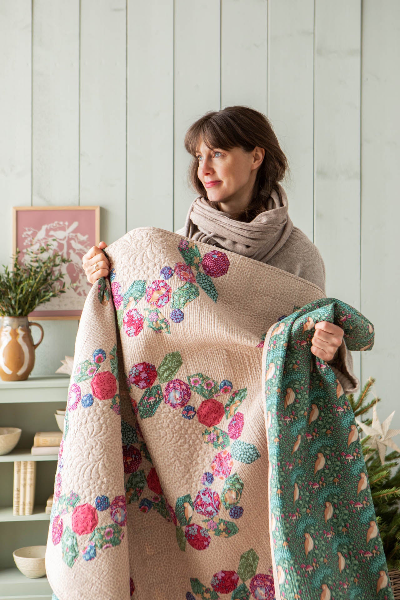 A woman wearing a beige knit sweater and scarf holds up a large quilt in a bright room. The blanket has a cream base with a colorful floral pattern in pink, red, and green, and shows a green reverse side with a small-scale floral print.
Photo by Tildafabrics.com [additional_image_link]