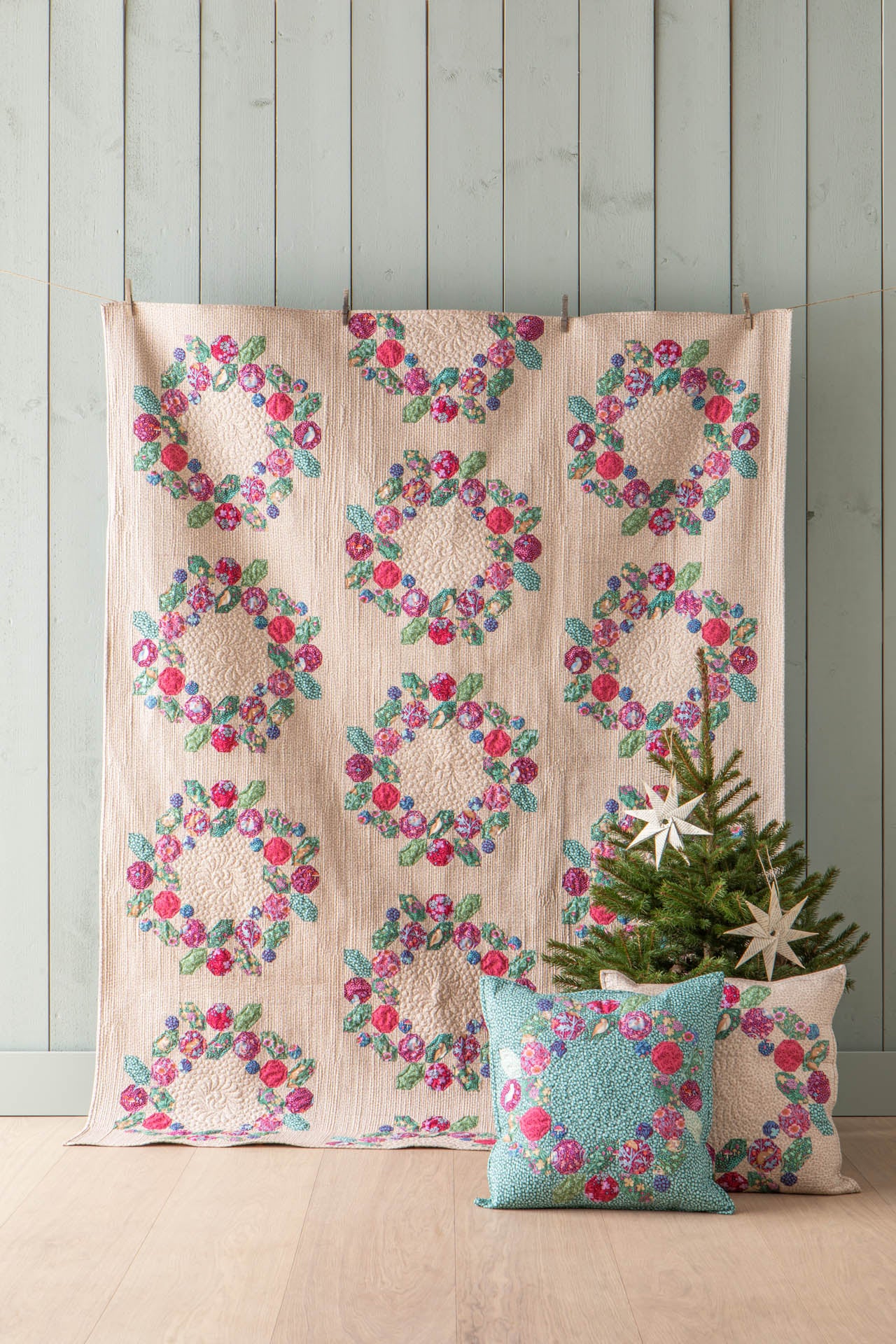 A quilted textile featuring repeating floral wreath motifs in pink, red, green, and blue on a light beige background, displayed against a pale wood-paneled wall with two matching decorative pillows and a small tree in front.
Photo by Tildafabrics.com [additional_image_link]