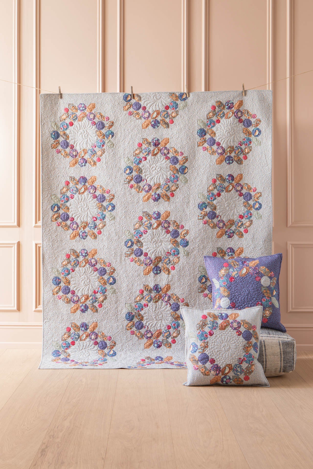 A quilted textile set displayed in a styled room: a large rectangular quilt hanging vertically and two matching square pillows in front, all featuring the same multicolored floral wreath pattern on a light background. The room has pale peach paneled walls and a light wood floor.
Photo by Tildafabrics.com [additional_image_link]
