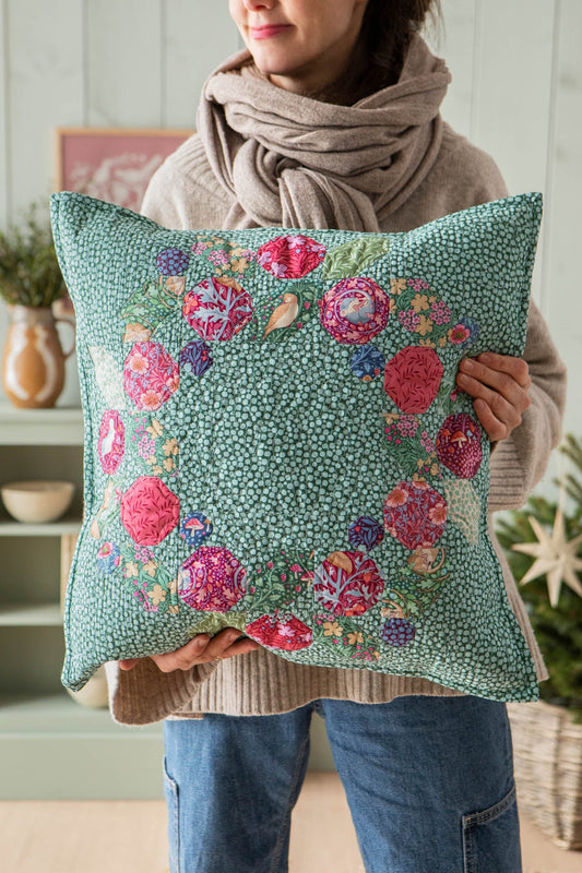 A person wearing a beige knit sweater and scarf holds a square decorative pillow. The pillow has a mint green background with a dense small floral pattern and a border of larger pink, red, purple, and blue flowers.
Photo by Tildafabrics.com [image_link]
