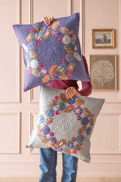 A person holds up two decorative square quilts in front of a pale pink paneled wall. The top quilt is lavender with a multicolor floral and paisley patchwork design, and the bottom quilt is light gray with a matching patchwork design.
Photo by Tildafabrics.com [image_link]