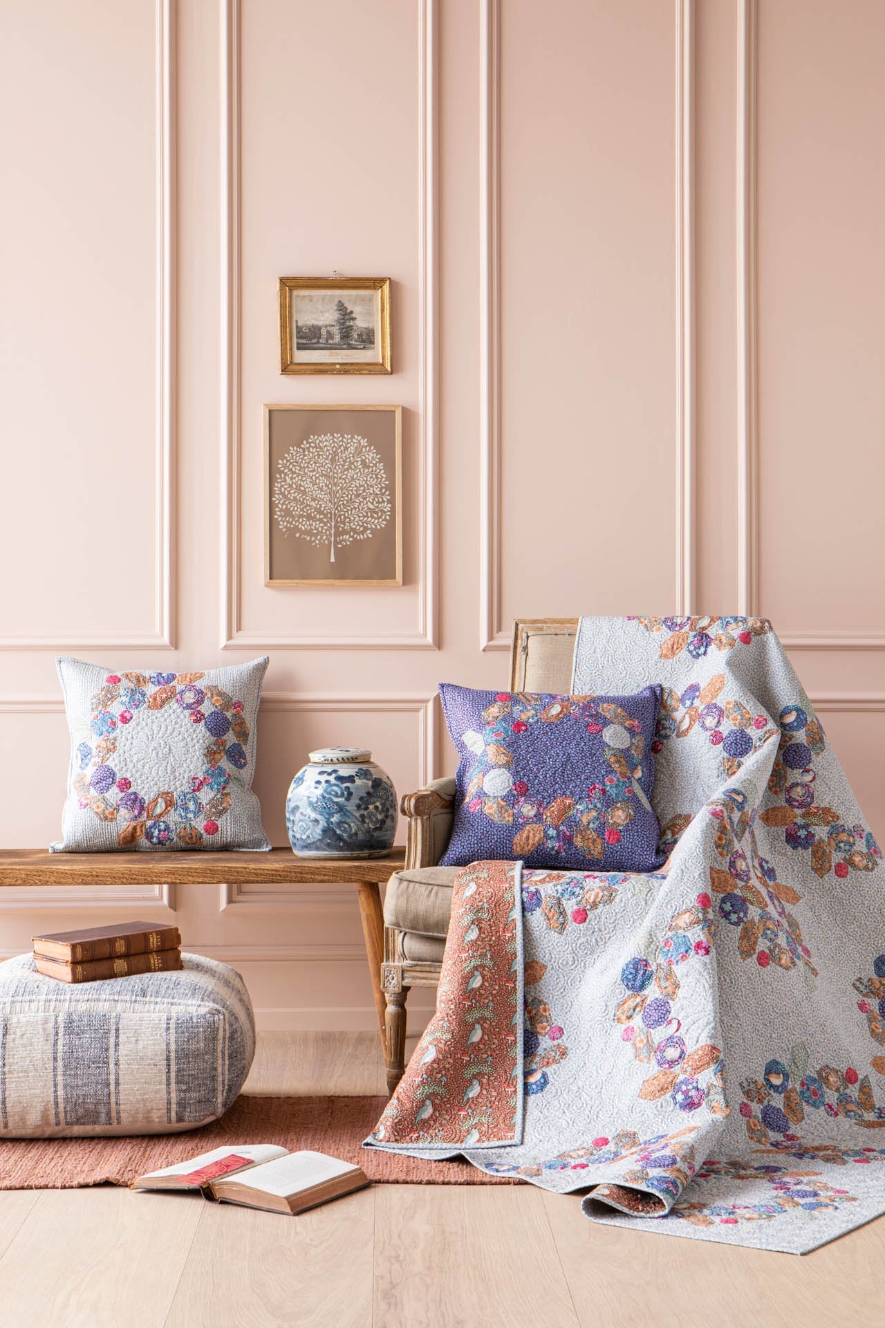 A styled interior scene with a floral quilt draped over a chair, matching patterned pillows, a bench, books, and a small vase against a pale pink paneled wall.
Photo by Tildafabrics.com [additional_image_link]