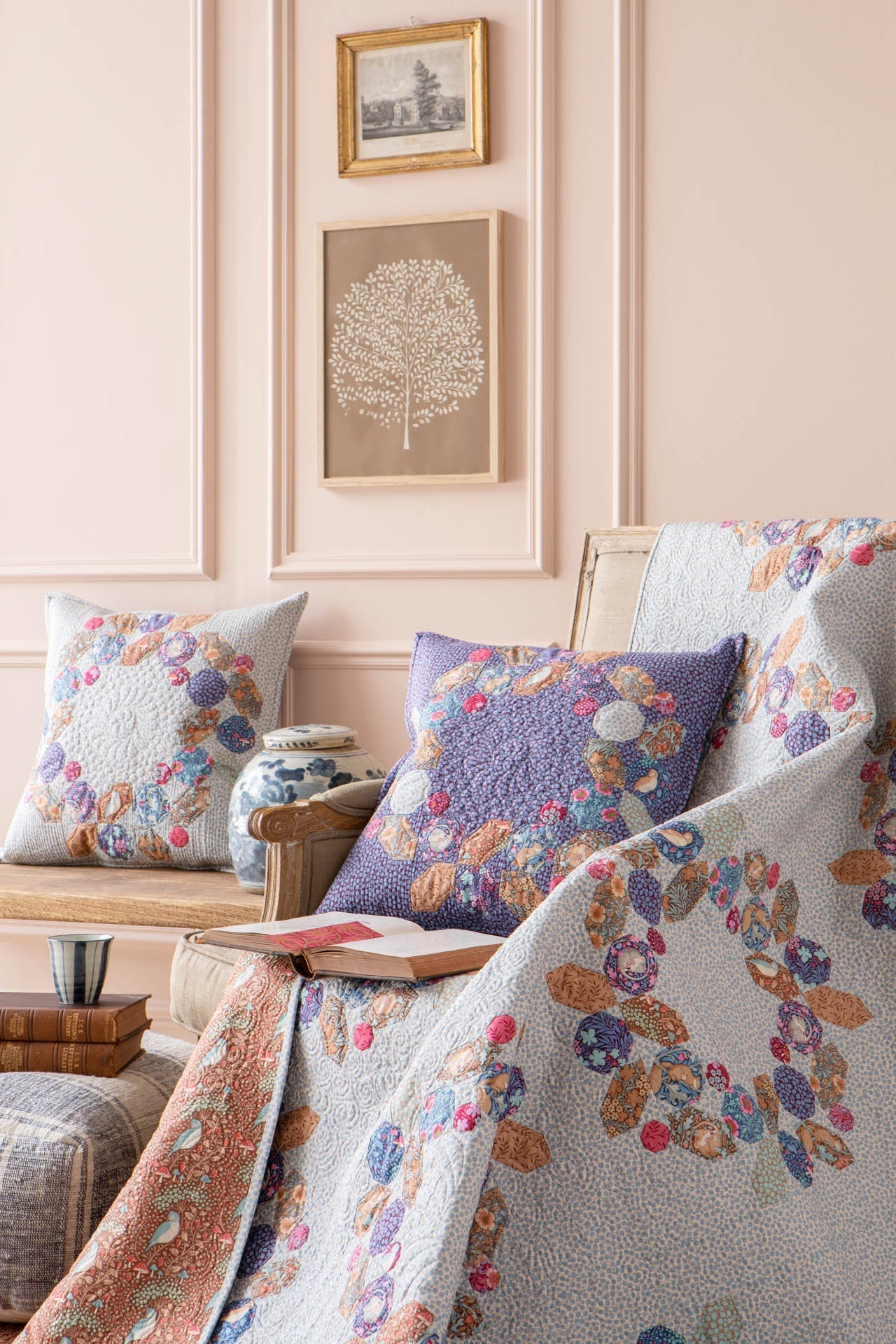 A cozy interior scene with a quilted throw blanket and matching pillows featuring a multicolor floral and botanical pattern, styled on a chair in a softly lit room with blush walls and framed wall art.
Photo by Tildafabrics.com [additional_image_link]