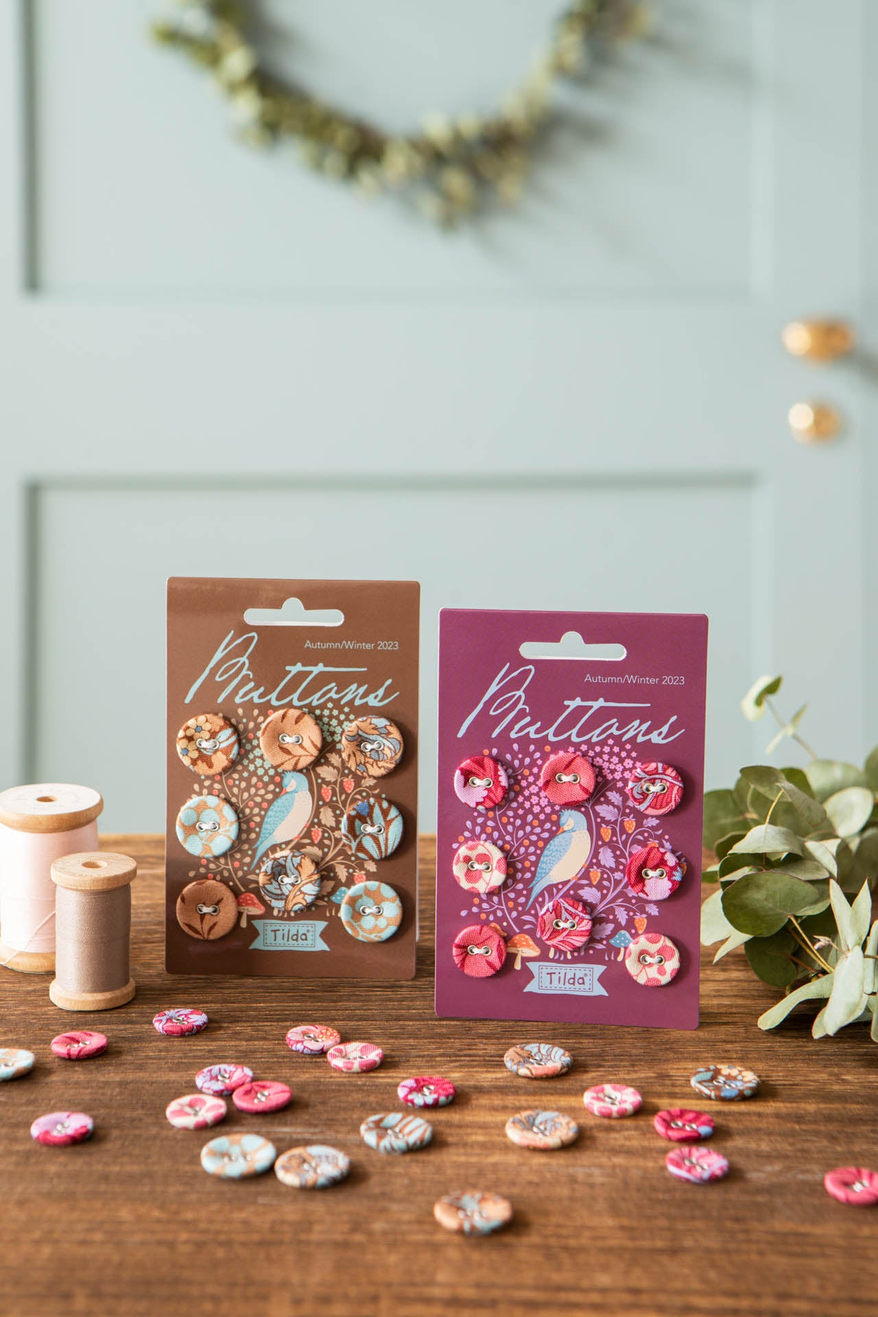 Two packages of Hibernation fabric buttons displayed on a wooden table, with loose buttons scattered around and sewing thread spools in the background.
Photo by Tildafabrics.com [additional_image_link]