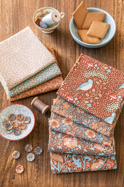 A styled flat-lay of multiple fabric bolts with intricate patterns including birds, foxes, and floral motifs, arranged on a rustic wooden table alongside sewing accessories like buttons, thread, and a teacup bowl of cookies.
Photo by Tildafabrics.com [additional_image_link]