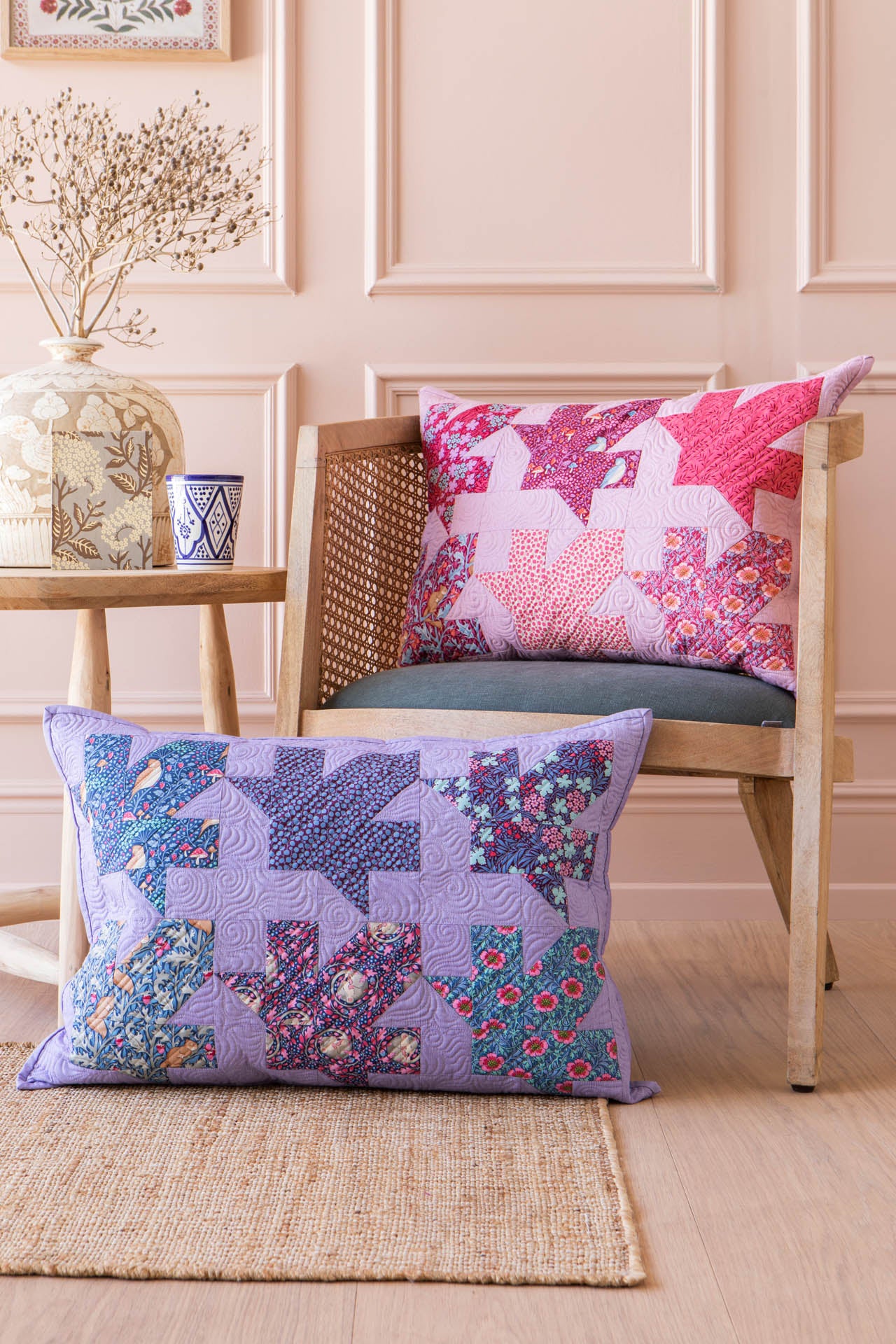 Two decorative patchwork lumbar pillows displayed in a styled room setting. The foreground pillow is lavender purple with blue and multicolor floral patchwork blocks, while the pillow on the wooden chair is pink and red with a star pattern.
Photo by Tildafabrics.com [image_link]