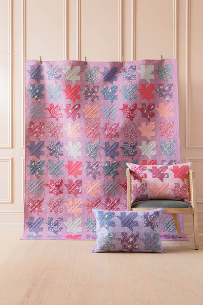 A pink patchwork quilt with coral, blue, and lavender geometric blocks is draped over a chair in a light wood-paneled room. A matching square throw pillow rests in front of the chair.
Photo by Tildafabrics.com [additional_image_link]