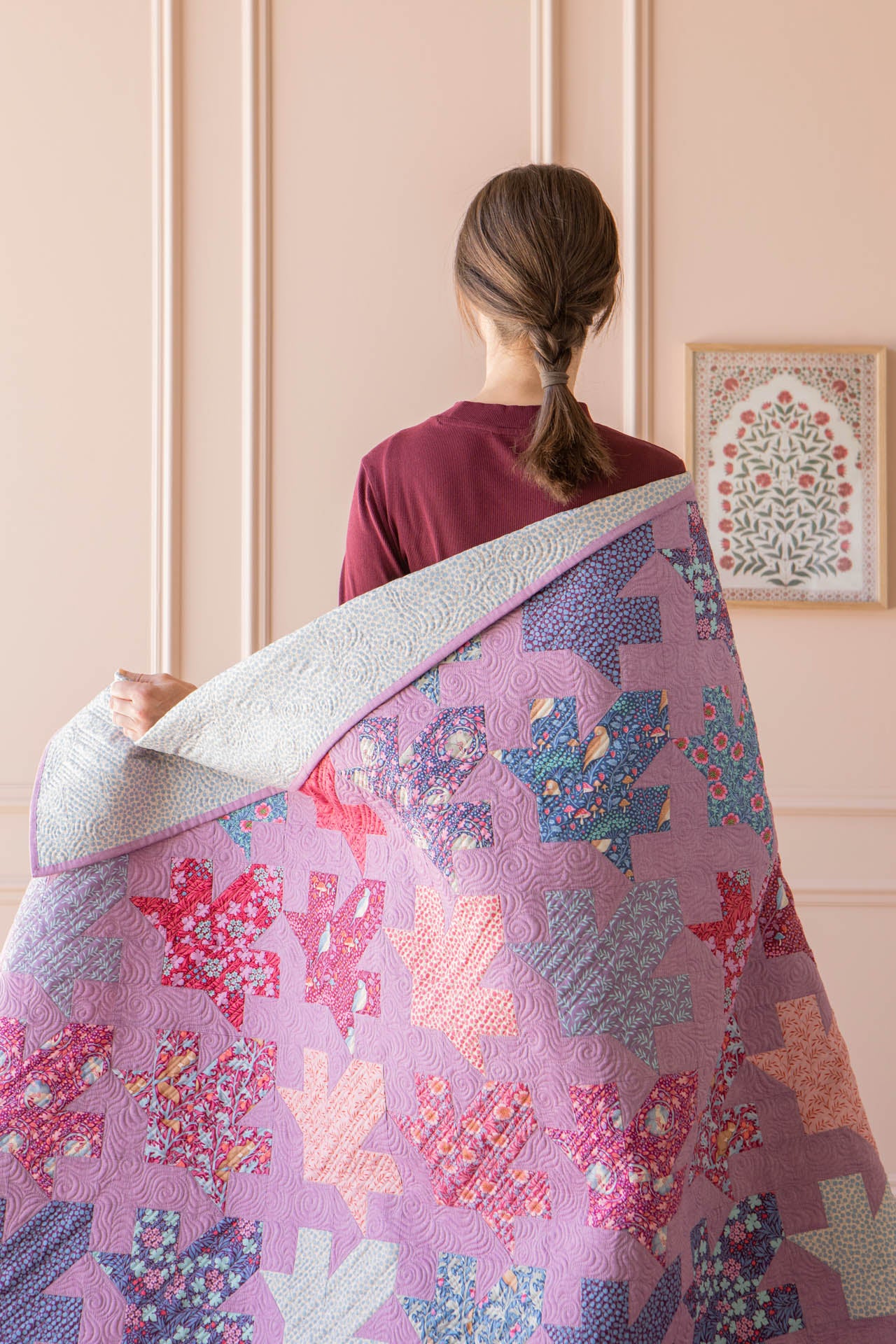 A woman viewed from behind holds up a large quilted blanket or throw in front of her. The quilt features a patchwork design with autumn leaf shapes in shades of pink, purple, blue, and orange, with a light gray reverse side.
Photo by Tildafabrics.com [additional_image_link]