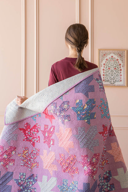 A woman viewed from behind holds up a large quilted blanket or throw in front of her. The quilt features a patchwork design with autumn leaf shapes in shades of pink, purple, blue, and orange, with a light gray reverse side.
Photo by Tildafabrics.com [additional_image_link]