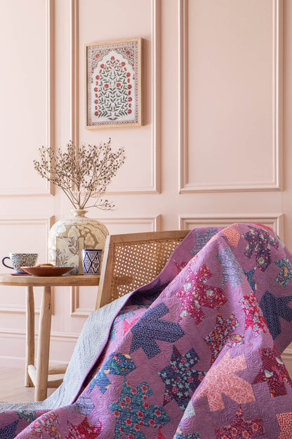 A purple and pink patchwork quilt with floral and geometric patterns draped over a wooden cane chair in a pink paneled room.
Photo by Tildafabrics.com [additional_image_link]