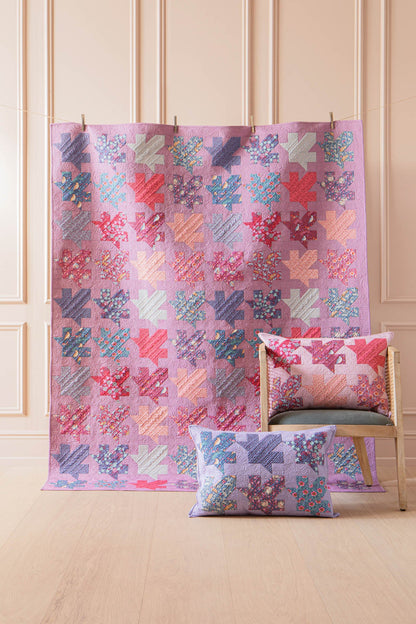 A pink patchwork quilt with coral, blue, and lavender geometric blocks is draped over a chair in a light wood-paneled room. A matching square throw pillow rests in front of the chair.
Photo by Tildafabrics.com [additional_image_link]
