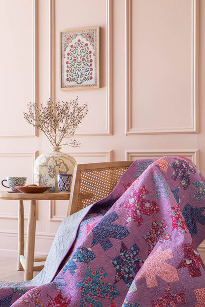 A purple and pink patchwork quilt with floral and geometric patterns draped over a wooden cane chair in a pink paneled room.
Photo by Tildafabrics.com [additional_image_link]