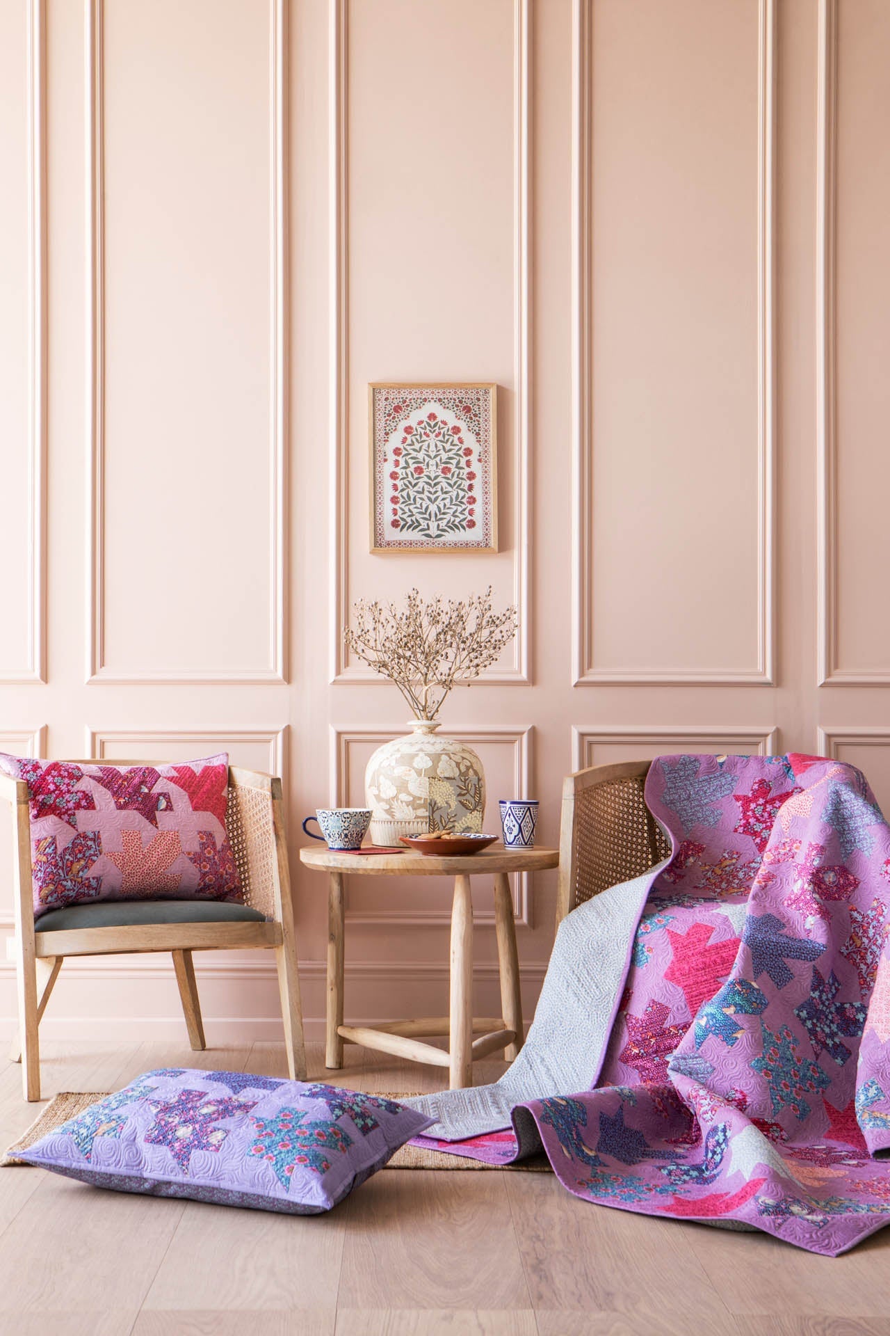 A styled interior scene with pink paneled walls, two wooden chairs, a small round table, and colorful patterned quilt and pillows draped over the chairs and floor. The quilt and pillows feature pink, purple, and blue floral motifs, with one pillow laid flat in the foreground.
Photo by Tildafabrics.com [additional_image_link]
