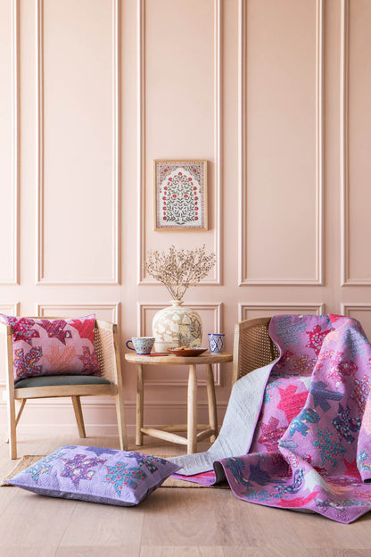 A styled interior scene with pink paneled walls, two wooden chairs, a small round table, and colorful patterned quilt and pillows draped over the chairs and floor. The quilt and pillows feature pink, purple, and blue floral motifs, with one pillow laid flat in the foreground.
Photo by Tildafabrics.com [additional_image_link]