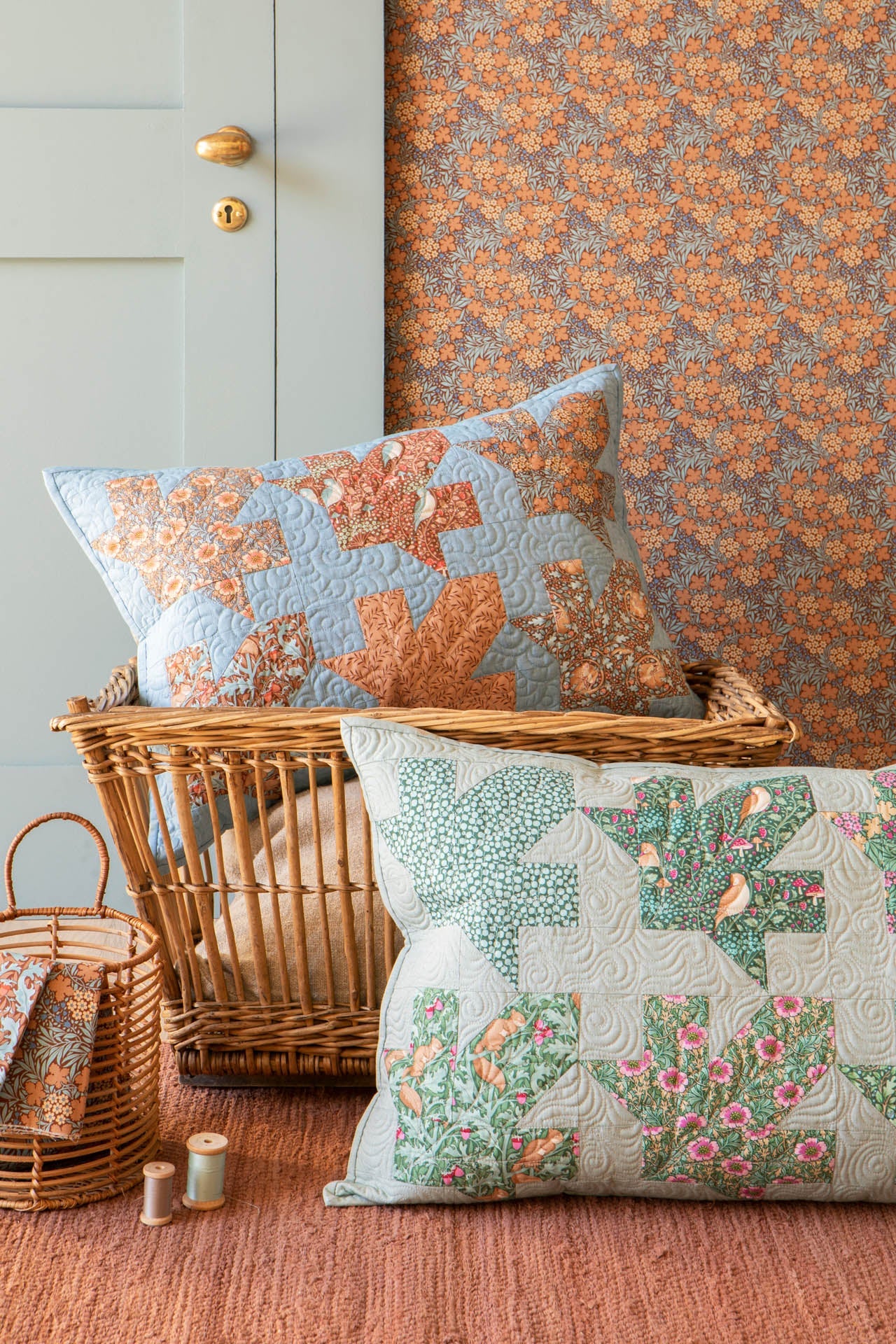 Two decorative throw pillows displayed in a styled interior setting. One pillow has a light blue background with orange and green floral/leaf patterns, and the other has a light gray background with green and pink floral quilted blocks.
Photo by Tildafabrics.com [image_link]