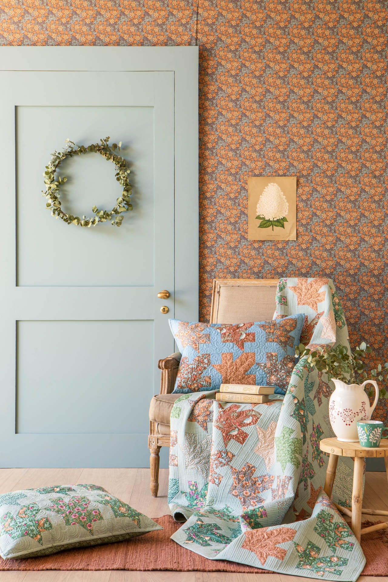 A cozy interior scene with a light blue paneled door, patterned wallpaper, a woven armchair draped with a floral quilt, matching pillows, a small side table with a pitcher, and a wreath on the door.
Photo by Tildafabrics.com [additional_image_link]