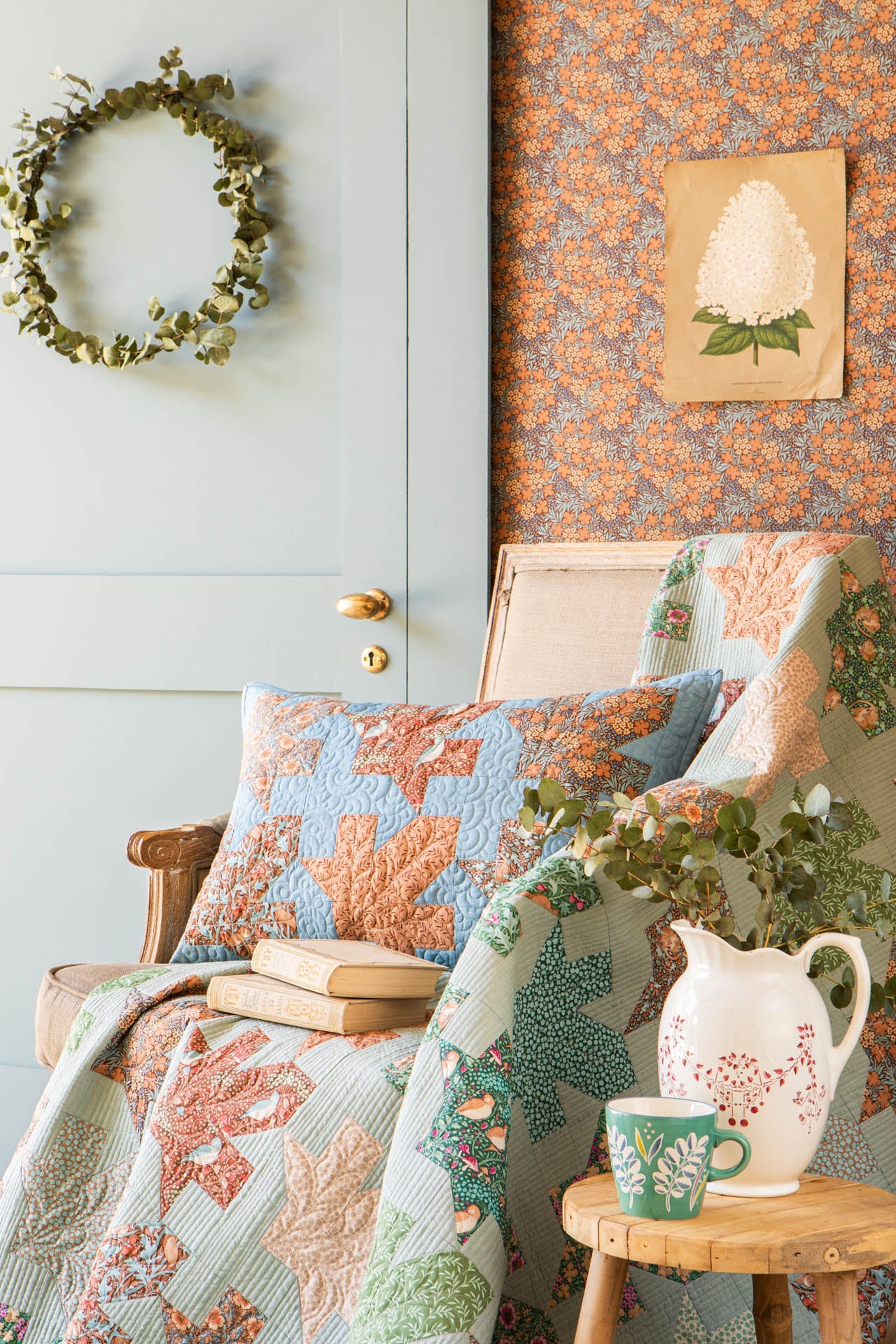 A cozy interior scene with a quilted throw blanket and matching pillow in soft blue, sage green, and peach tones, styled on a chair beside a small wooden side table with a white pitcher, green mug, and books. A pale blue paneled door, floral wallpaper, and a green wreath are visible in the background.
Photo by Tildafabrics.com [additional_image_link]