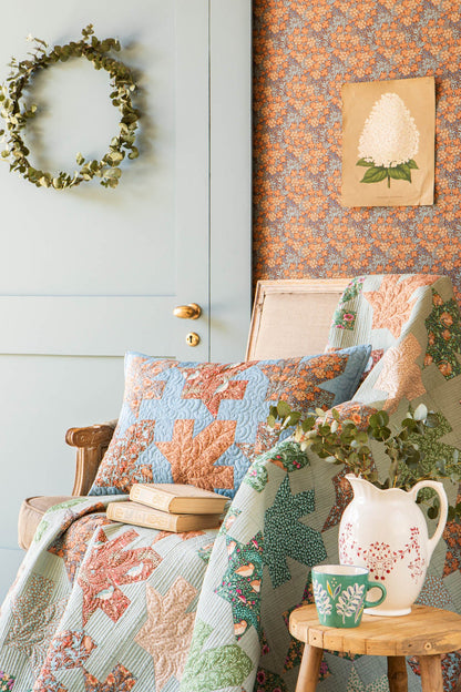 A cozy interior scene with a quilted throw blanket and matching pillow in soft blue, sage green, and peach tones, styled on a chair beside a small wooden side table with a white pitcher, green mug, and books. A pale blue paneled door, floral wallpaper, and a green wreath are visible in the background.
Photo by Tildafabrics.com [additional_image_link]