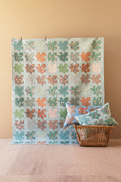 A quilted patchwork textile with repeating coral and sea-creature motifs in muted orange, teal, sage green, and cream, displayed hanging against a warm beige wall above a light wood floor. A wicker basket holds two matching decorative pillows.
Photo by Tildafabrics.com [additional_image_link]