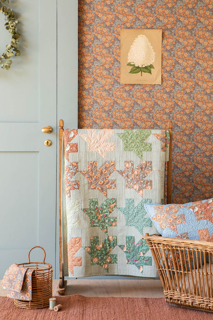 A quilted textile with a patchwork leaf pattern in muted green, peach, and blue is draped over a wooden stand in a styled room. The background features patterned wallpaper, a light blue paneled door, a small woven basket, and a wicker chair with a matching pillow.
Photo by Tildafabrics.com [additional_image_link]