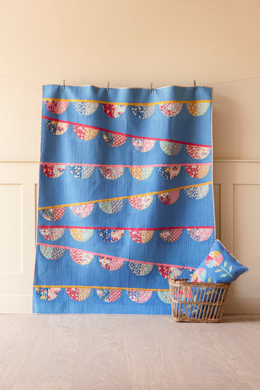 A blue fabric wall hanging or curtain with repeating scalloped bunting patterns in pink, red, yellow, and cream, displayed on a string in a room with a beige wall and light wood floor. A matching blue pillow and a wicker basket are placed beside it.
Photo by Tildafabrics.com [image_link]