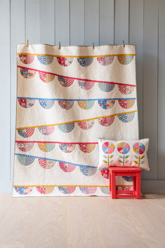A cream-colored quilt with colorful, patterned scalloped stripes hangs against a light gray paneled wall, with a matching decorative pillow resting on a small red stool.
Photo by Tildafabrics.com [image_link]