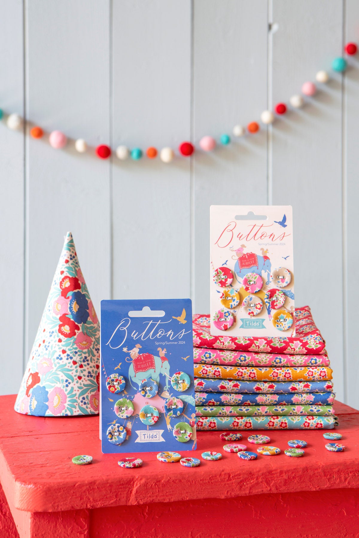 A styled product display of floral buttons and matching fabric. The set includes a cone hat, a stack of folded patterned fabric, a blue card of buttons, a white card of buttons, and several loose buttons scattered on a red surface against a light gray paneled wall with a colorful pom-pom garland.
Photo by Tildafabrics.com [additional_image_link]