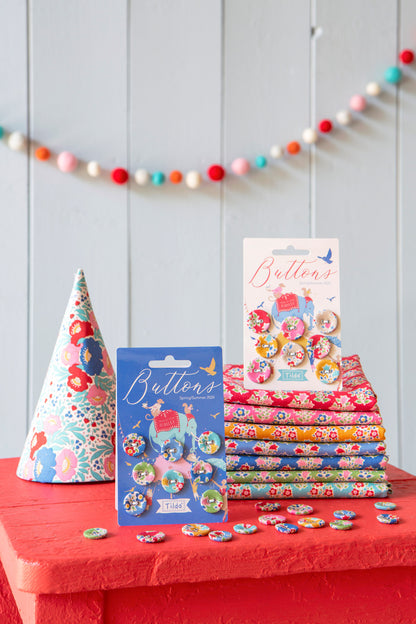 A styled product display of floral buttons and matching fabric. The set includes a cone hat, a stack of folded patterned fabric, a blue card of buttons, a white card of buttons, and several loose buttons scattered on a red surface against a light gray paneled wall with a colorful pom-pom garland.
Photo by Tildafabrics.com [additional_image_link]